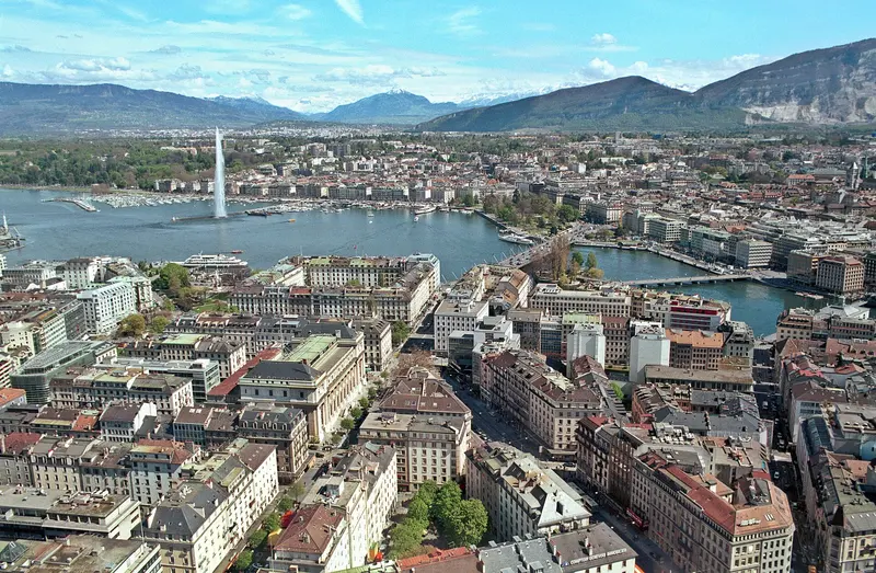 View over the city of Geneva and its lake with a tall fountain and mountains behind